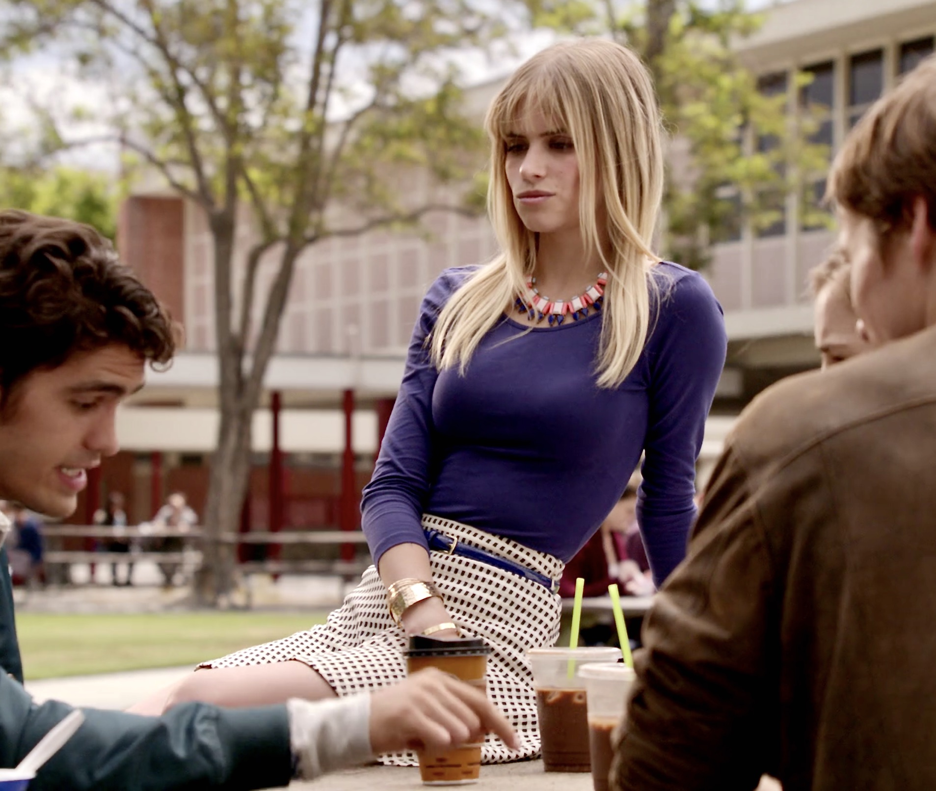 Brooke Maddox sitting on top of an outdoor lunch table. She is wearing a purple top, a white-black checked mini skirt, and a multi-colored statement necklace..